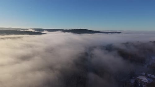 Aerial view of a dense layer of fog drifting over forested hills under a clear blue sky. Soft