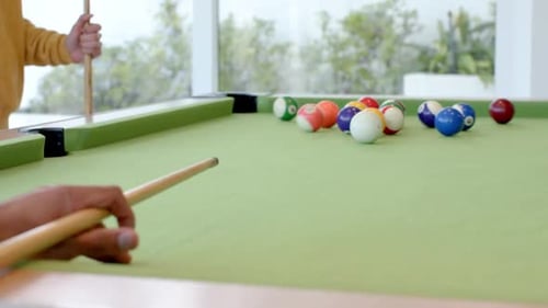 O jogador está colocando uma bola branca, alinhando o bastão e batendo para quebrar bolas coloridas na mesa de sinuca