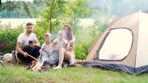 camping by the lake in the woods. happy family dad mom and little kids sitting by the fire and tent
