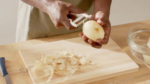Man wearing green apron peeling potato with white Y-shaped peeler on wooden board, preparing dinner
