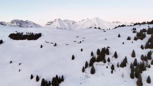 Aerial view of a snow-covered mountain landscape at dusk with evergreen trees and isolated cabins
