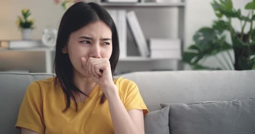 Sick young Asian woman coughing while sitting on sofa at living room