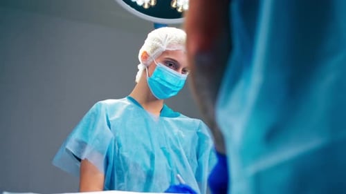 A girl assistant surgeon in uniform and a medical mask passes scalpel to her colleague an operation