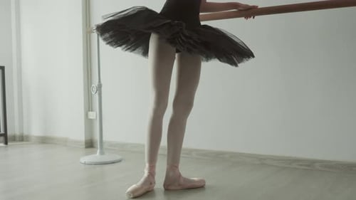 Dancer in black tutu practices at barre, showcasing elegance and poise in softly lit studio.