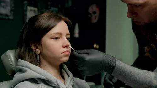 Interior de un estudio de piercings: el maestro de piercings hace un piercing para una niña, piercing para el tabique nasal