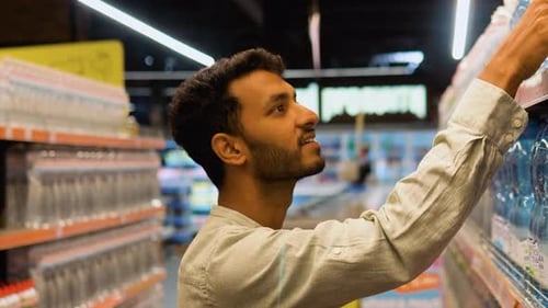 Handsome Indian Guy Holding Shopping Basket and Buying Mineral Water at Supermarket