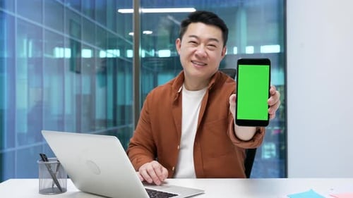 Asian businessman presenting mobile phone with green screen, sitting at desk in business office.
