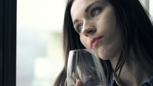 Pensive woman drinking red wine by window indoors