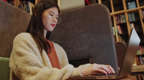 Asian Female Student Typing on Laptop in Modern Library