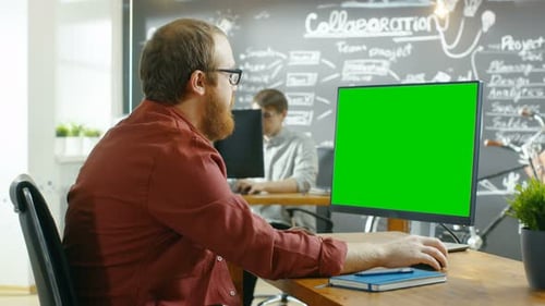 Man Works on the Personal Computer with Green Mock-up Screen. In the background Creative Office wit