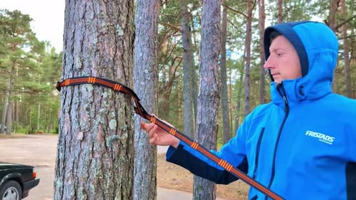 Un homme resserre la sangle d'un hamac dans une forêt, capturant le moment de l'installation d'un camping relaxant