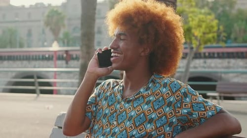 Side-view portrait of young man having phone talk sitting outdoors and smiling