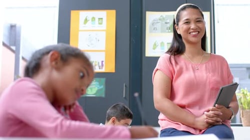 Smiling teacher holding tablet in classroom, students writing in school