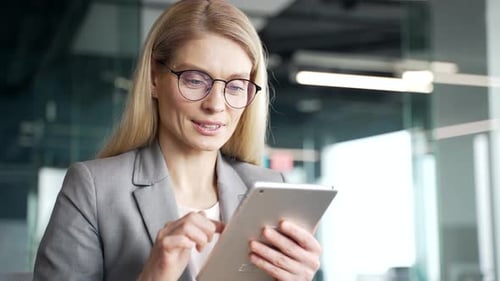 Confident businesswoman in a jacket is using digital tablet standing in business office. Smiling