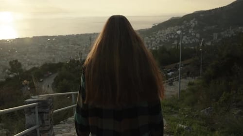 Following a young woman descending a stone path from the mountain, with the city below.