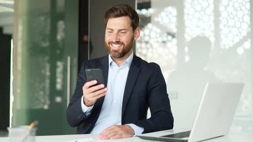 Smiling businessman is using smartphone while sitting at workplace in business office. Worker reads