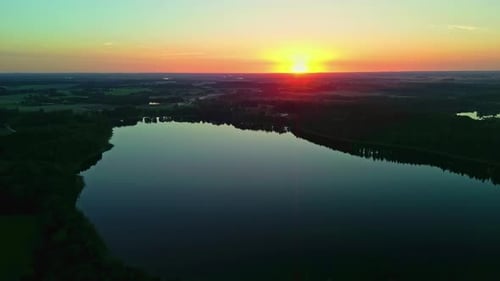 Aerial shot flying backward revealing a serene lake at sunset surrounded by trees with vibrant sky c