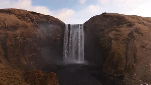 Slow drone push-in toward Skógafoss waterfall in Iceland - misty cliffs and rugged golden landscape