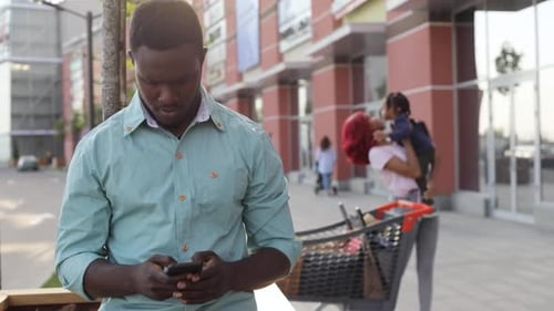 Handsome AfroAmerican Man Uses His Smartphone By the Entrance to Shopping Center While His Cheerful