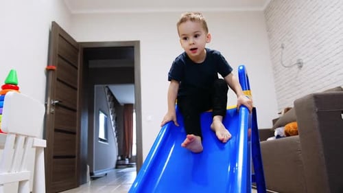 Caucasian baby in blue t-shirt and black pants climbing by the plastic slide indoors.