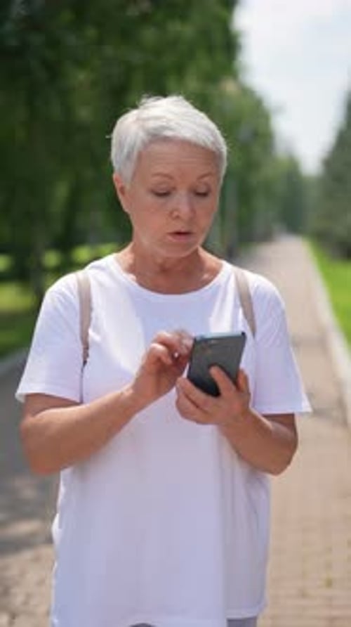 Vertical Portrait of Senior Aged Woman Typing on Smartphone While Walking in City Park Beautiful