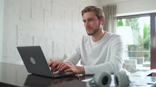 Handheld Focused Man in Wireless Headphones Sits at Home on the Desk and Works Using a Laptop Trader
