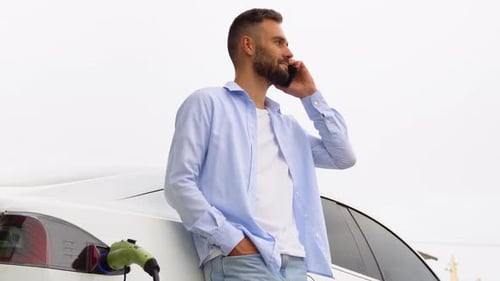 Stylish Man Phoning While Charging Car at Electric Vehicle Charging Station