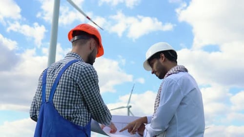 Indian and Caucasian Workers Discussing a Blueprint on a Wind Farm in Front of a Turbine Supplying