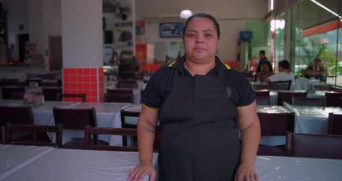 Latina restaurant employee standing in dining area, neutral expression, wearing apron in modern