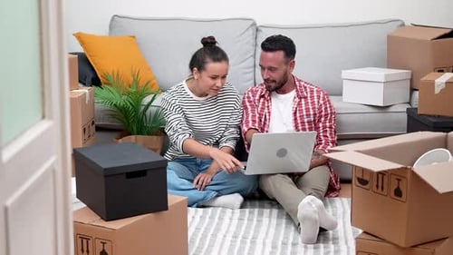 Young Couple in New Home Amid Boxes Uses Laptop to Decide on Interior Design Shop Online