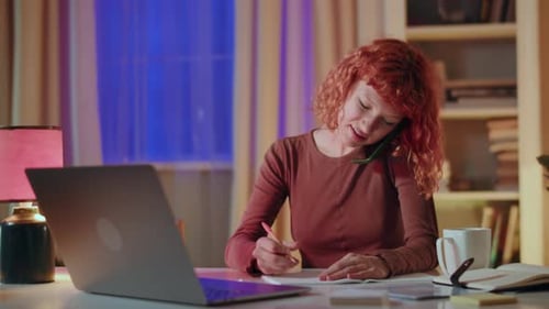 A Young Woman with Red Hair is Focused on Her Work Multitasking on Her Laptop and Taking Notes