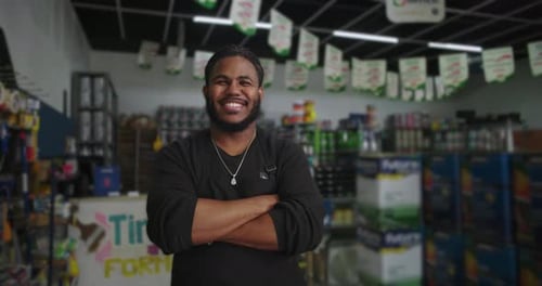 Man in black apron smiling confidently with arms crossed in a paint store, surrounded by shelves of