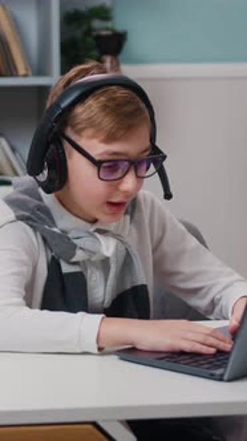 Fröhlicher kleiner Junge mit Brille und Kopfhörern spielt Videospiel mit Laptop, der an seinem sitzt