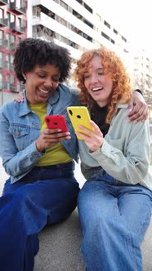 Two Smiling Women Look at Smartphones in the City