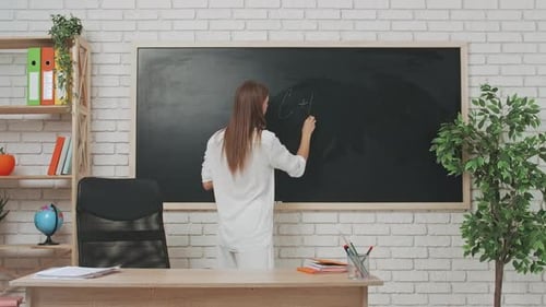 Young Woman College Teacher at Desk in Classroom in Front of Chalkboard Explains Chemistry Lesson to