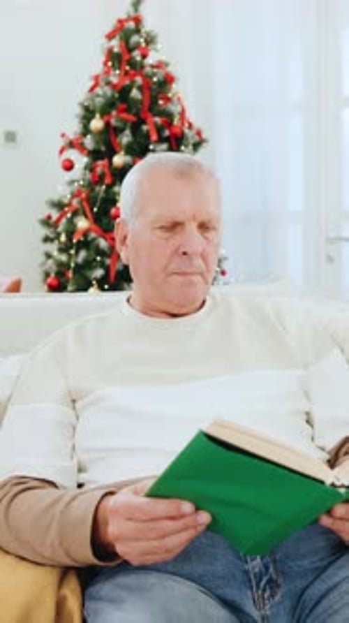 Elderly Man Reading a Book By the Christmas Tree While His Family Enjoys New Year Celebrations in a