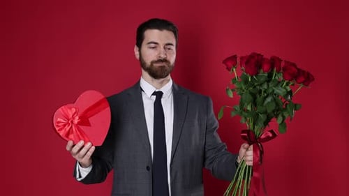 St. Valentine's Day. Handsome man with bouquet of roses and gift on red background