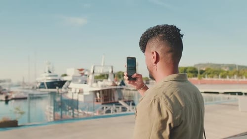 Young man stands in the port, looks at yachts, takes photos of yachts and ships