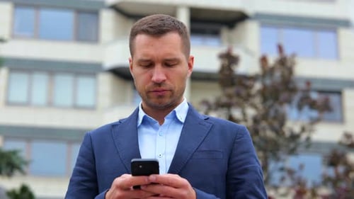 Handsome Caucasian Stylish Man with Serious Face Standing in Street in Town Texting on Smartphone