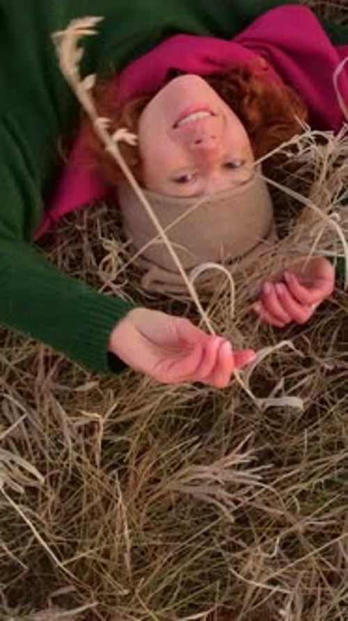 Woman Lying in Dry Grass Smiling and Holding Strands of Grass Wearing Warm Clothing and a Hat
