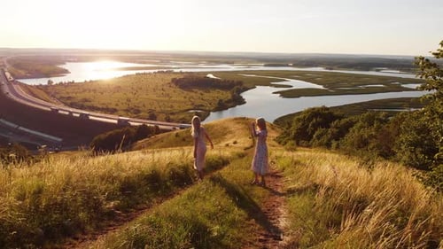 Two Young Blonde Women in Summer Light Dresses Holding Hands Walking Down the Hill Above the Highway
