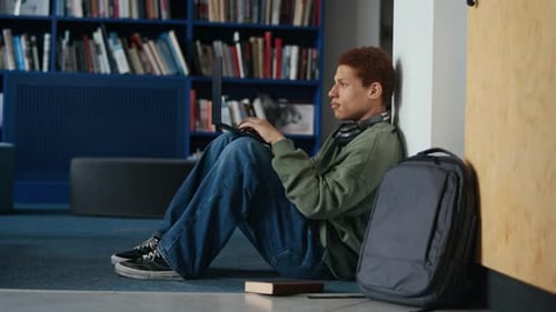 Estudiante masculino birracial concentrado que usa una computadora portátil sentado en el piso de la biblioteca