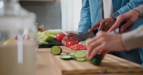 Close up shot. Male hands slicing a tomatoes and female hands cutting cucumber on a wooden cutting b