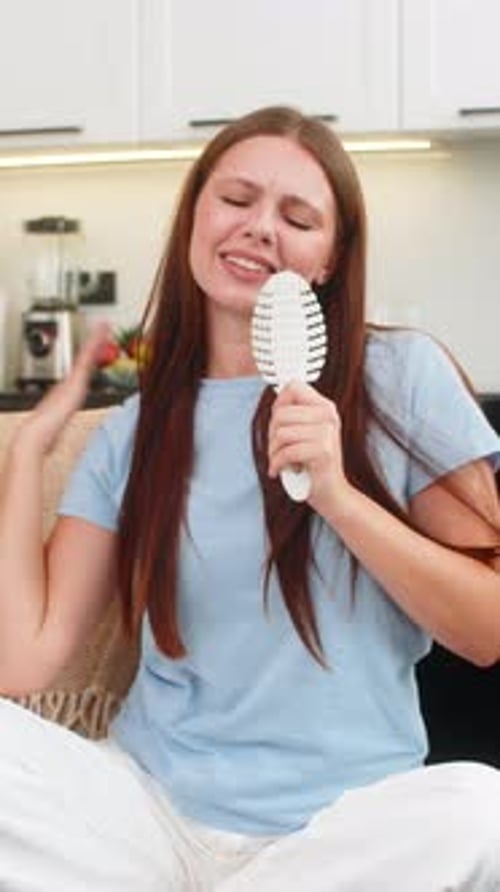 Happy Woman Singing into Hairbrush on Sofa at Home