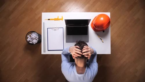 Engineer at Cluttered White Desk Clutching Head Beside Laptop Hard Hat Crumpled Papers and Clipboard