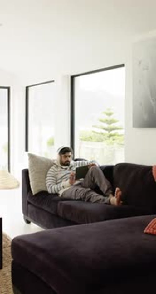 Vidéo verticale : homme indien allongé sur un canapé, tapotant une tablette avec un casque dans un salon lumineux