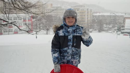 Happy smiling boy holding his plastic sleds on a snowy hill at snowfall