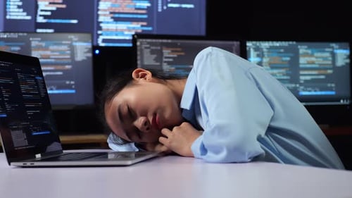 Vue latérale d'une programmeuse asiatique endormie pendant qu'elle écrit du code sur un ordinateur portable utilisant plusieurs moniteurs