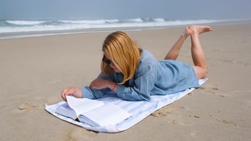 Eine junge Frau liegt auf dem Bauch auf einem Strandtuch und liest ein Buch am Atlantik, das sie genießt