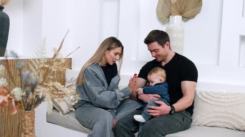 Les parents caucasiens sont à la maison avec leur bébé. L'enfant entre les mains de papa est concentré sur un bracelet.